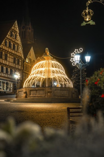 Illuminated fountain in front of half-timbered houses surrounded by festive Christmas lights at night, Calw, Black Forest, Germany