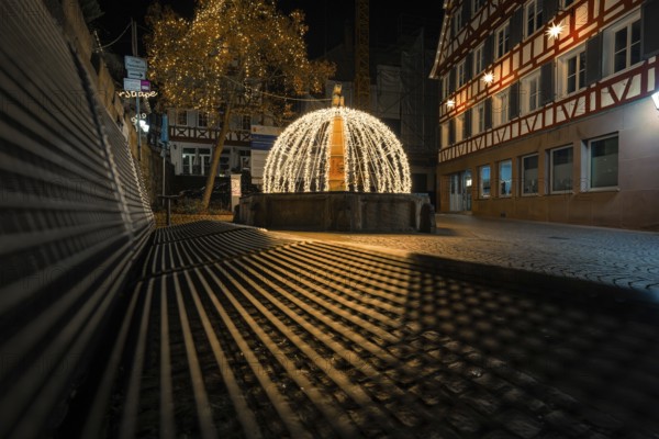 Night view of a festively illuminated fountain with shadows cast in a historic street, Calw, Black Forest, Germany