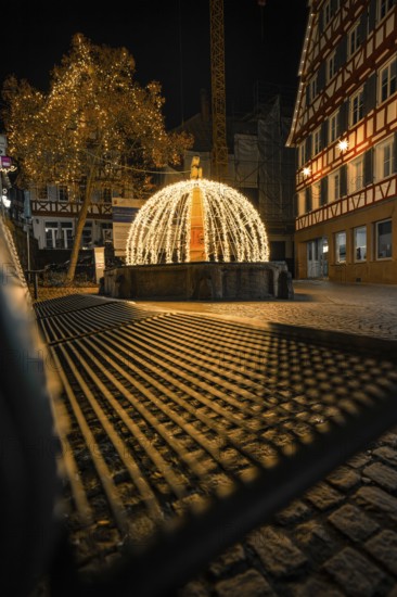 Festively illuminated fountain with lights and shadows on a paved road at night, Calw, Black Forest, Germany
