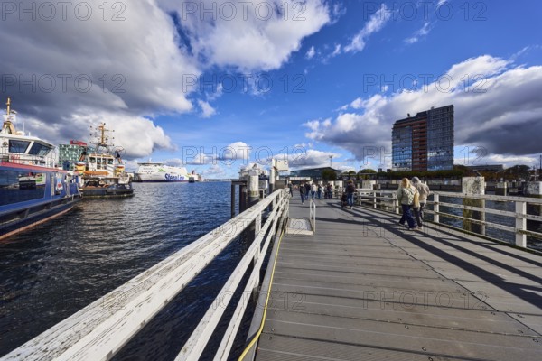 Kiel Fjord, station bridge, pier, wooden railings, wharf, ferries, harbor and sea tugs, ships, high-rise center at Germania port, pedestrians as accessories, blue sky, cumulus clouds, stratocumulus clouds, white and dark clouds, Kiel, state capital, district-free city, Schleswig-Holstein, Germany