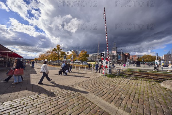 Pedestrian crossing with barrier, St. Andrew's cross, track bed, tracks, general architecture, trees, autumn leaves, sidewalk made of concrete paving stones and cobblestones, pedestrians as accessories, shadow, side light, blue sky, cumulus clouds, stratocumulus clouds, white and dark clouds, intersection of station bridge, quay road and Kiel sailor square, Kiel, state capital, district-free city, Schleswig-Holstein, Germany