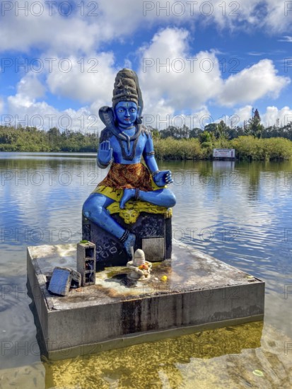 Statue sculpture of deity Shiva on the shore of stands in sacred Hindu lake Ganga Talao at religious site largest Hindu sanctuary for religion Hinduism outside India for believing Hindu Hindus, in the background Sacred Lake Ganga Talao crater lake in crater of extinct volcano, Grand Bassin, Le Pétrin, Mauritius