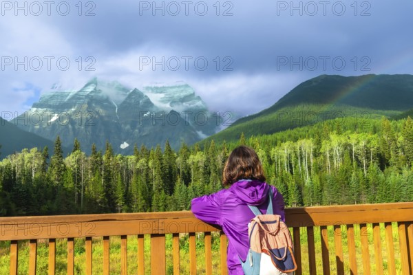 Female hiker with a backpack enjoying the breathtaking view of mount robson, the highest peak in the canadian rockies, beneath a vibrant rainbow over lush forests