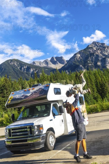 Father lifting his son in the air, celebrating their adventure in mount robson provincial park, british columbia, with their camper van parked nearby under a bright blue sky