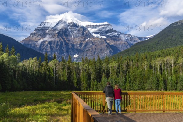 Two tourists standing on a wooden deck are enjoying the breathtaking view of mount robson, the highest peak in the canadian rockies, surrounded by lush green forest and a vibrant meadow