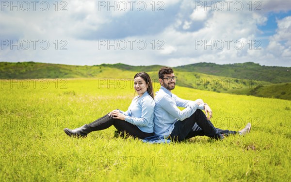 Couple in love in the field sitting back to back looking at the the camera, Young relaxed couple sitting in the field with their backs to each other lon the field