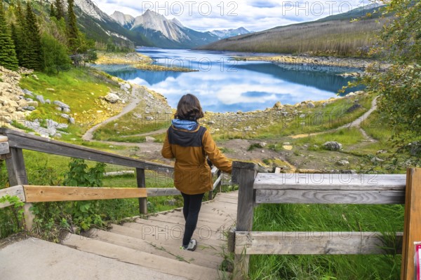 Female tourist walking down wooden stairs to medicine lake, with turquoise waters reflecting clouds, surrounded by mountains and forests in jasper national park, alberta
