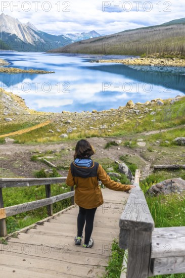 Female tourist walking down wooden stairs approaching medicine lake with turquoise water reflecting clouds and mountains in the background, located in jasper national park, alberta, canada