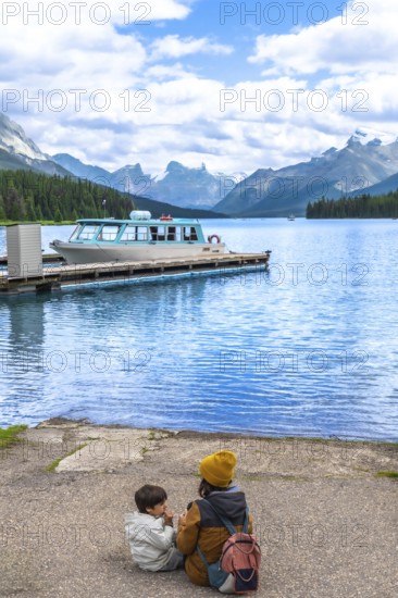 Mother and son sitting on maligne lake's shore, admiring the turquoise water and a tour boat at the dock, with spirit island and the canadian rockies in view