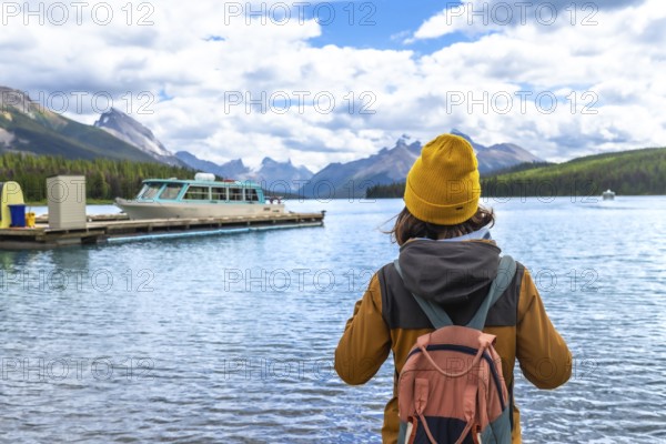Female tourist wearing a yellow beanie and carrying a backpack admires the stunning view of maligne lake and spirit island in jasper national park, alberta
