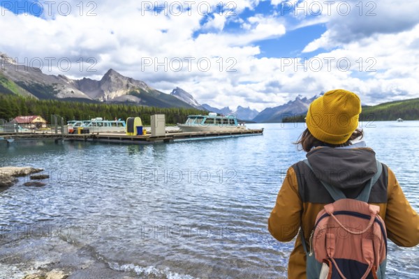 Female tourist wearing a yellow beanie and carrying a backpack admires the stunning landscape of maligne lake with spirit island and boats in the background