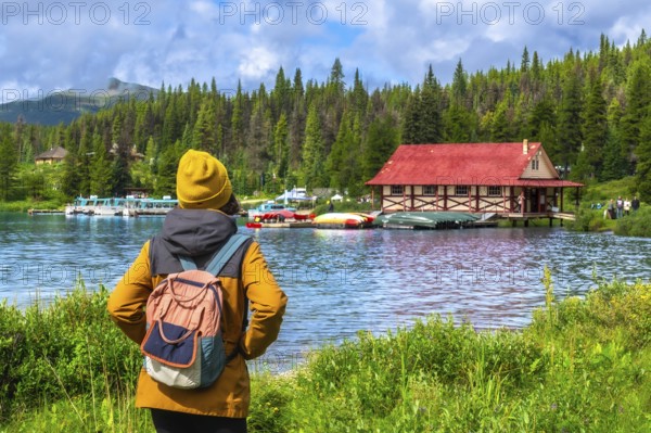 Female tourist wearing a yellow beanie and backpack admiring the iconic boathouse at maligne lake in jasper national park, surrounded by stunning nature on a summer day