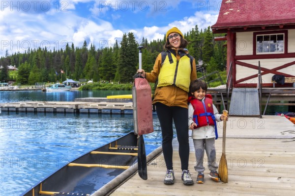 Mother and son holding paddles and wearing life jackets, standing on a wooden dock beside a canoe at maligne lake in jasper national park, alberta, canada