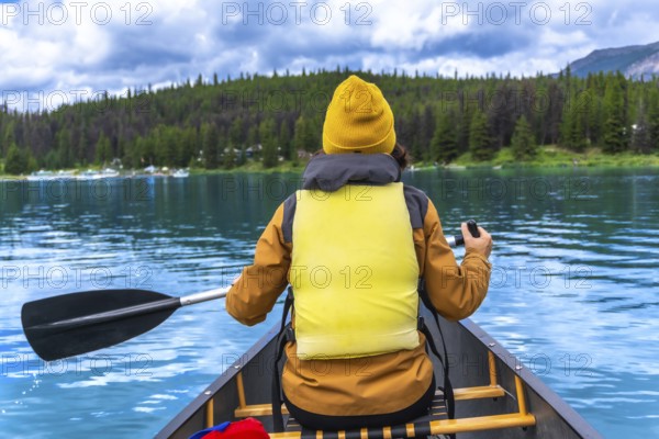 Tourist wearing yellow life jacket and beanie paddling a canoe in maligne lake, jasper national park, alberta, enjoying the turquoise water and mountain view on a cloudy summer day