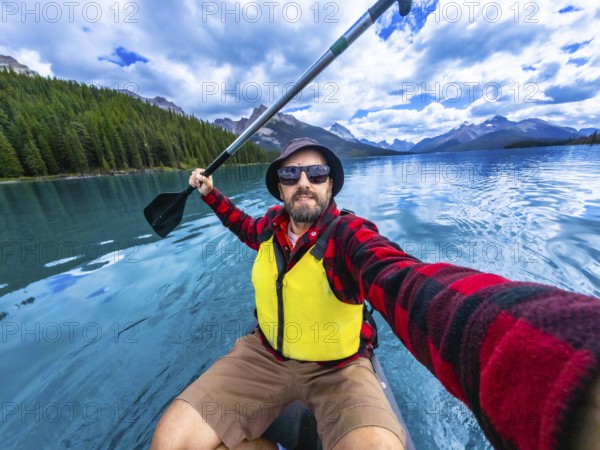 Tourist wearing sunglasses and life jacket kayaking and taking a selfie in maligne lake with beautiful mountain range reflection in the turquoise water in jasper national park, alberta, canada