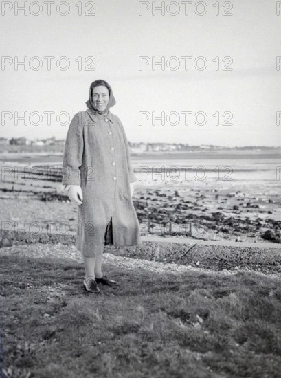Full length portrait of smiling woman wearing winter coat and clothing standing by seaside, Devon, England, UKL c 1956