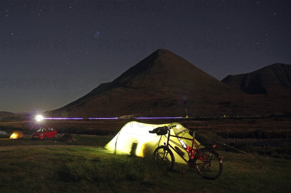 An illuminated tent next to a bicycle in front of a mountain on a clear starry night, Isle of Skye, Hebrides, Scotland, United Kingdom