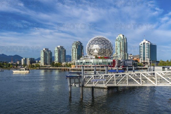 Modern buildings and science world are reflecting in the calm waters of false creek, vancouver, with mountains in the background under a beautiful blue sky on a sunny day