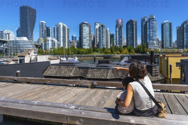 Mother and son sitting on a wooden boardwalk, pointing towards the stunning vancouver skyline from granville island market on a sunny day