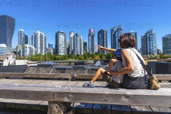 Tourist mother pointing at the skyline of vancouver while sitting with her son on a wooden bench at granville island market in british columbia, canada, during a sunny summer day