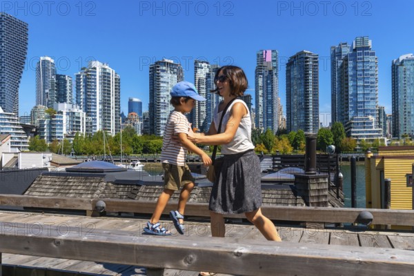 Mother and son enjoying a sunny day walking along the wooden boardwalk on granville island with the cityscape of vancouver, british columbia, canada, in the background