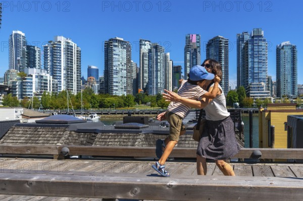 Mother and her son are sharing a tender hug on a wooden pier in vancouver's yaletown neighborhood, with the city's modern skyline and waterfront serving as a backdrop on a sunny day
