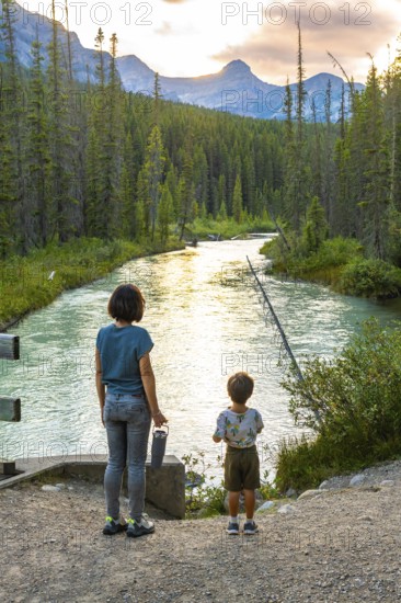 Mother and her son are enjoying the breathtaking view of a pristine river flowing through a lush forest, surrounded by the majestic canadian rockies at sunset in banff national park, alberta