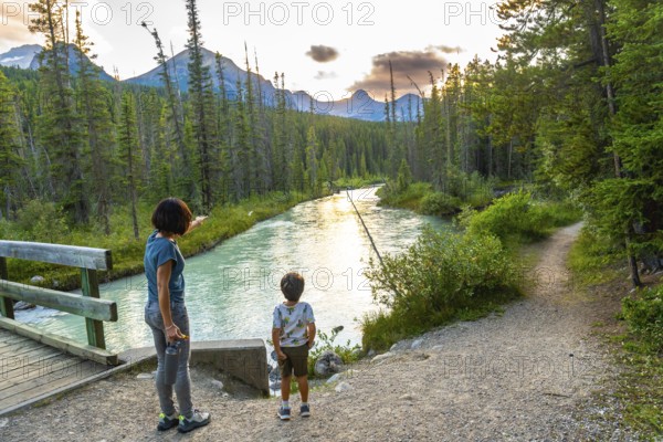 Woman points towards the majestic mountains in the distance as she and her son stand near a tranquil river in banff national park, enjoying the serene sunset view
