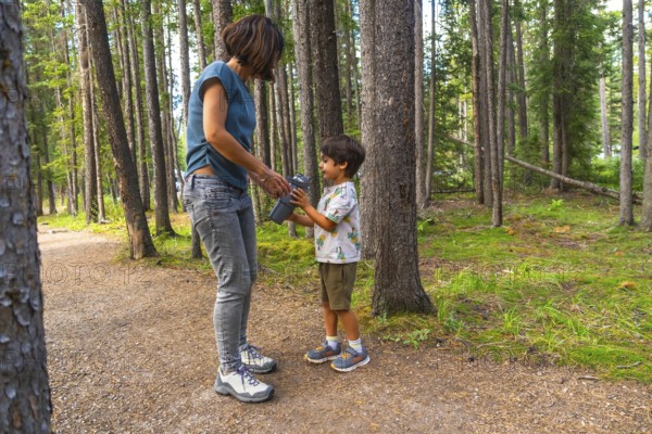 Mother hands a water bottle to her son during a hike on a sunny day surrounded by the lush green trees of banff national park in the canadian rockies