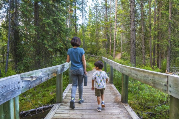 Mother and son enjoying a leisurely hike, crossing a picturesque wooden bridge amidst the lush green forest of banff national park in the canadian rockies, creating cherished memories together