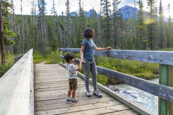 Mother and son using binoculars observing the landscape from a wooden footbridge over a creek in banff national park, canadian rockies, alberta, canada