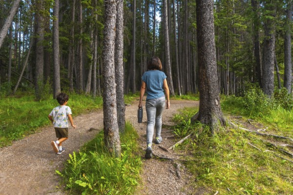 Mother carrying a water bottle and her son enjoying a leisurely hike along a picturesque forest trail in banff national park, surrounded by lush greenery and towering trees