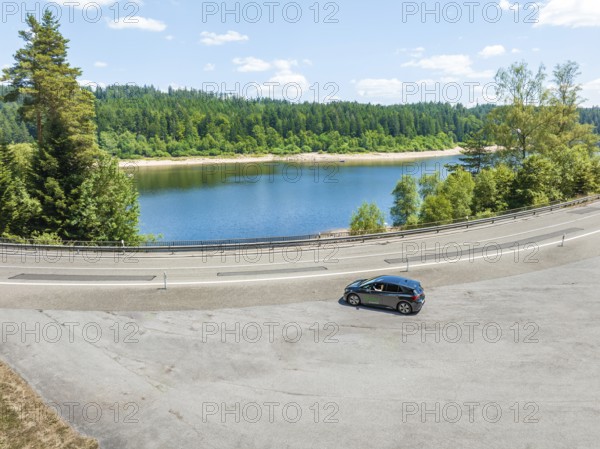 A black car drives along a lake, surrounded by a forest, in clear skies, deer e-car sharing, Cupra electric car, Black Forest, Germany