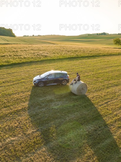 Car with person on a hay bale in a wide field at sunset, deer e-car sharing, Cupra electric car, Gechingen, Germany