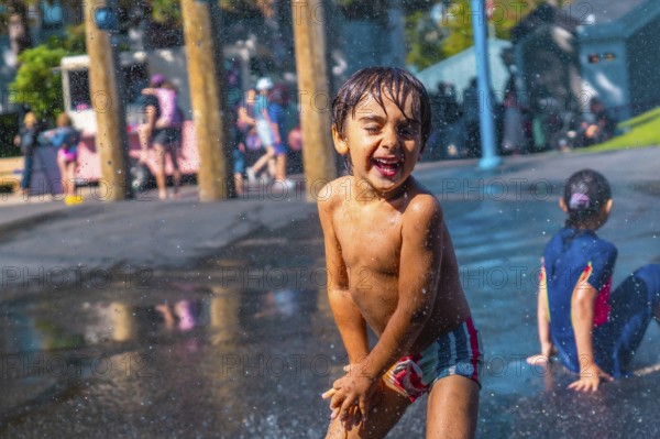 Cheerful child with wet hair and colorful swimming trunks laughing and playing in a city park splash pad, enjoying refreshing water sprays on a sunny day