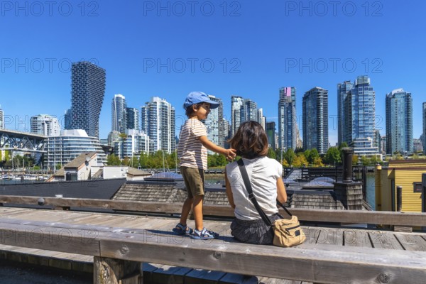 Sitting on a wooden bench at granville island market, a mother and son admire the stunning vancouver skyline under a clear blue sky on a sunny day