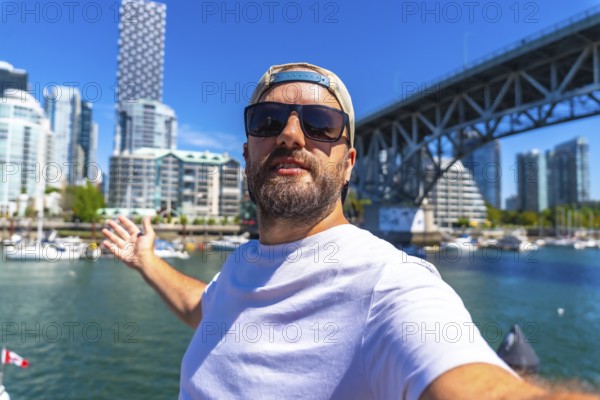 Happy man taking selfie with outstretched arm showing vancouver skyline, cambie street bridge and false creek with boats in british columbia, canada, during a sunny summer day