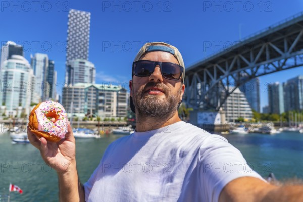 Tourist taking a selfie while holding a donut with burrard bridge and the cityscape of vancouver in the background, during a sunny summer day