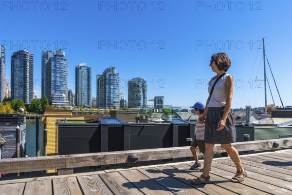Mother and her son enjoying a sunny summer day walking along the wooden boardwalk of granville island market with the cityscape of vancouver in the background