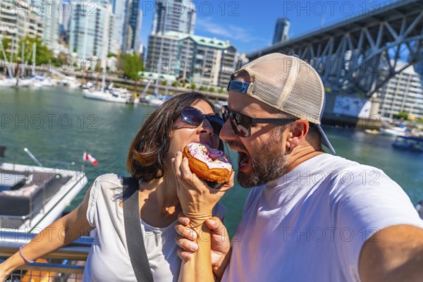 Happy couple biting into a donut together, taking a selfie with vancouver's cityscape and burrard bridge in the background, enjoying a sweet treat during their vacation