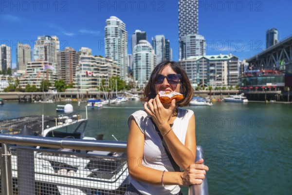Young woman enjoying a delicious donut with the stunning cityscape of vancouver, british columbia, as her backdrop, showcasing a perfect blend of urban exploration and sweet indulgence