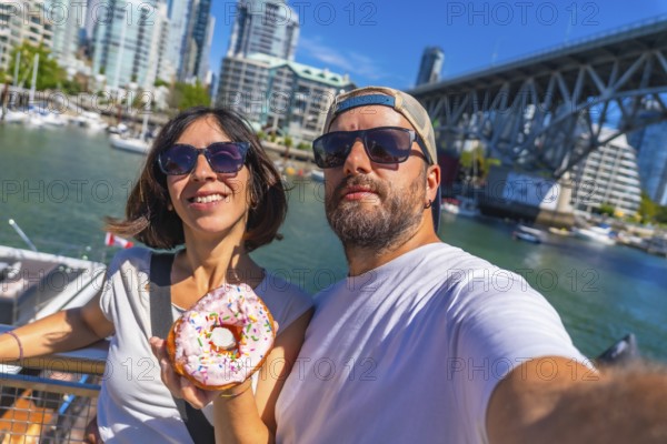 Happy tourists taking a selfie while holding a donut with sprinkles in front of the cityscape and burrard bridge in vancouver harbor, british columbia, canada