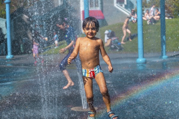 Happy child playing and running under the water jets of a splash pad in vancouver, british columbia, during a sunny summer day, creating a rainbow effect