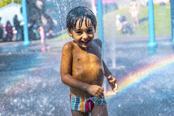 Happy child running and playing under splash pad jets with rainbow in the background on a hot summer day at a water park, enjoying the refreshing spray and having fun