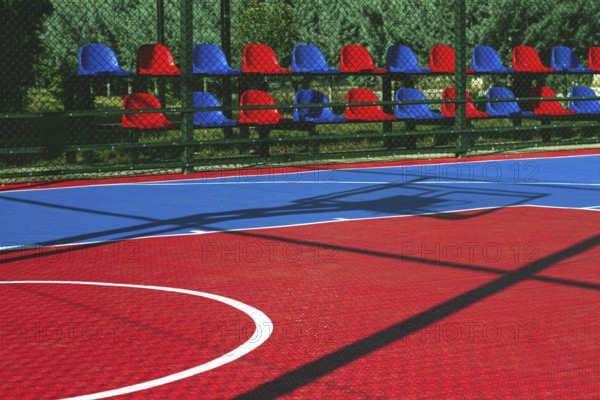 A vibrant basketball court features a mix of red and blue colors. Empty seats line the side, indicating a lively atmosphere earlier in the day. The sunlight casts interesting shadows on the surface