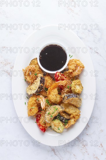 Shrimp and assorted vegetables are deep fried in a light batter and served on a white plate. A small bowl of soy sauce is beside the dish