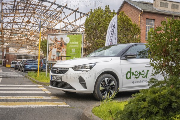 An Opel with car sharing logo stands in an urban environment with billboard and vegetation, Deer E-Car Sharing, Motorworld Rüsselsheim, Germany