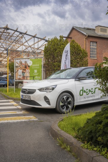 Opel with car sharing logo stands in front of an advertising banner in an urban setting with vegetation, Deer E-Car Sharing, Motorworld Rüsselsheim, Germany