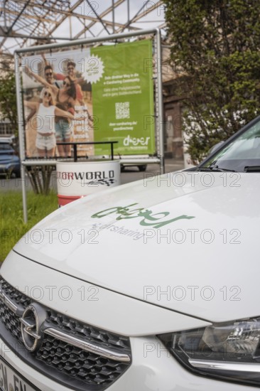 A white Opel with car sharing logo stands in front of an advertising poster and green surroundings, Deer E-Car Sharing, Motorworld Rüsselsheim, Germany