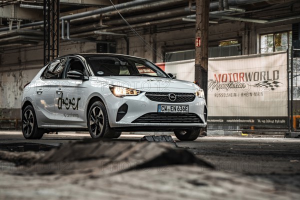 A white car is parked in front of a Motorworld banner in an industrial hall, Deer E-Car Sharing, Motorworld Rüsselsheim, Germany